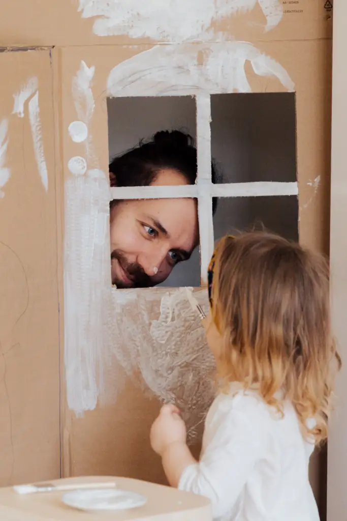 Father peeking through a cardboard playhouse window at a young child.